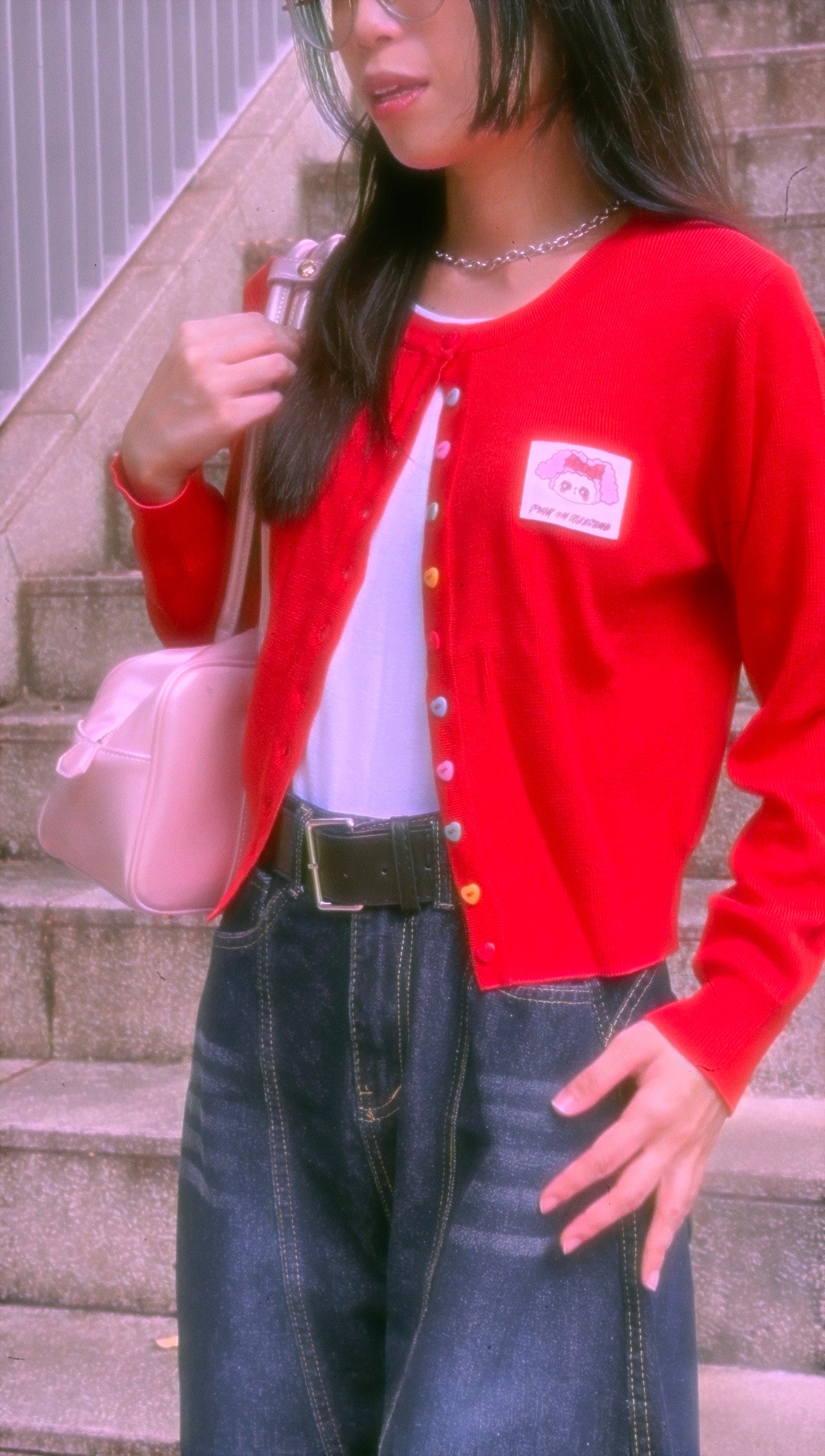 Person wearing a red cardigan with a logo, white shirt, and blue jeans, holding a pink bag.